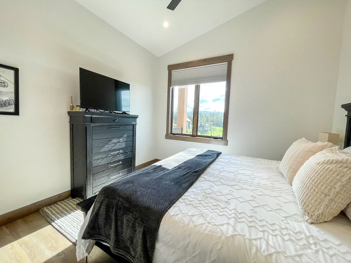 A serene bedroom featuring a bed with crisp white bedding, a large window offering scenic views, and a black dresser topped with a 50" Smart TV. The setting is inviting and tranquil, ideal for restful nights and relaxing movie sessions.