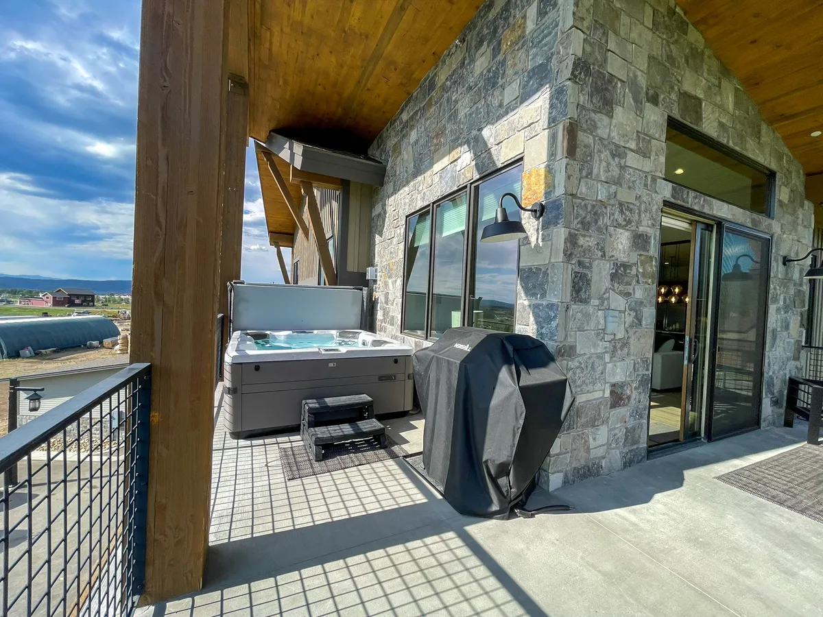 An inviting outdoor living area showcases a bubbling hot tub, an outdoor grill set against a stylish stone wall, and large glass doors connecting interior comforts to the exterior. The space is ideal for relaxing soaks, entertaining guests, and enjoying beautiful views.
