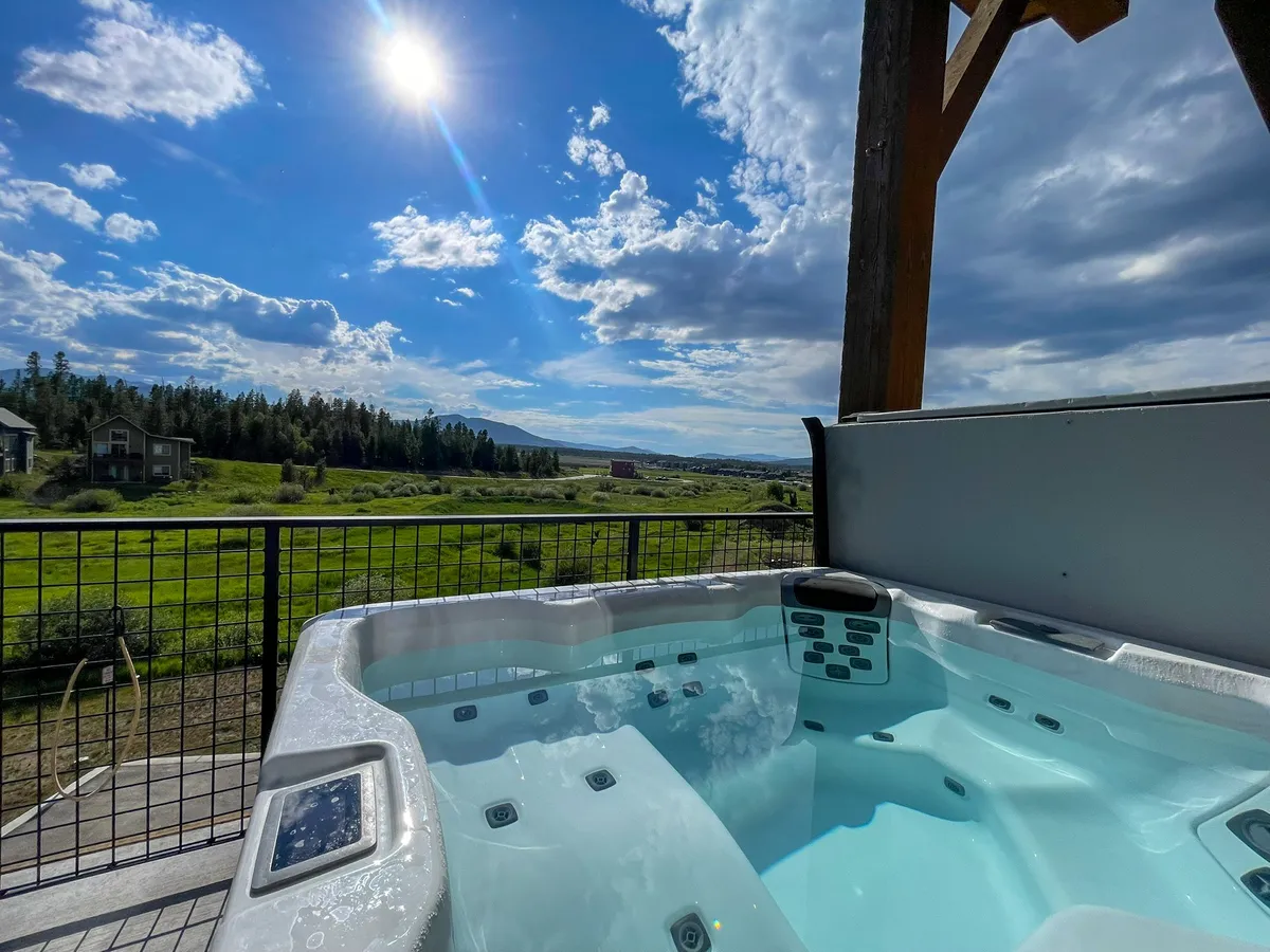 An elevated deck featuring a bubbling hot tub with seating, overlooking a stunning mountain panorama under open skies. The mood is tranquil yet inviting, hinting at après-adventure relaxation.