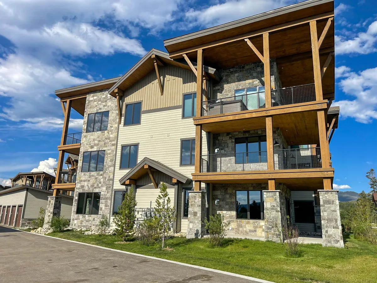 A wide exterior view of the condo’s stone and wood architecture, featuring balconies with railings and well-maintained landscaping. The overall look is modern yet welcoming, with ample outdoor space for gathering. The backdrop suggests proximity to nature and seasonal activities.