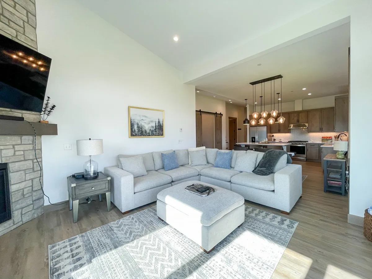 A spacious open-concept living area featuring a large sectional sofa, a cozy fireplace, and a view into the adjacent kitchen area. Chandelier lighting adds warmth, and bright daylight streams through large windows, highlighting both the living and kitchen spaces.