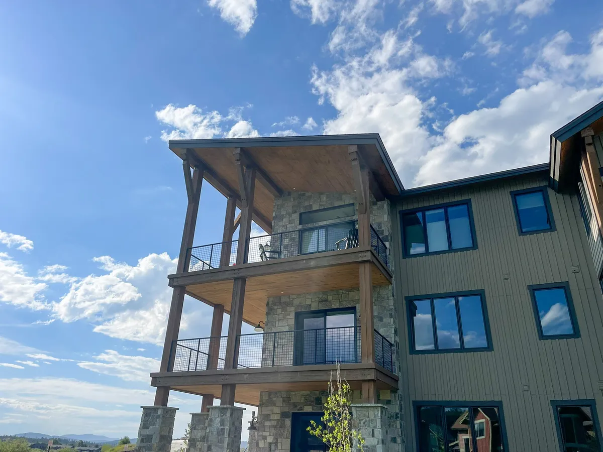 A close-up of a balcony with wooden beams and a stone facade, set against a blue sky. The balcony invites outdoor relaxation and offers clear views of the surrounding peaks, ideal for enjoying morning coffee or evening downtime. The space looks serene and perfectly integrated with the mountain environment.
