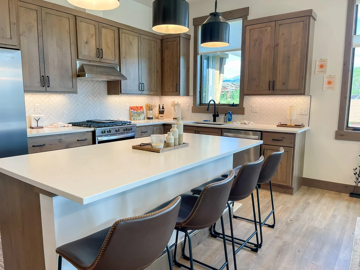 The image showcases a stunning, open-concept kitchen featuring a spacious kitchen island, gleaming stainless steel appliances, rich wood cabinetry, and a stylish herringbone backsplash. The space is bathed in warm lighting, creating an inviting atmosphere perfect for cooking, entertaining, and gathering with loved ones.